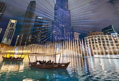 Panoramic view of the Dubai Fountain at night with Abras in the foreground and Burj Khalifa and its laser show in the background.